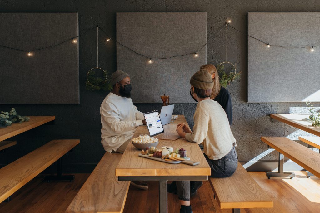 Man with hat and mask talks to woman in cream sweater facing him, both with laptops open